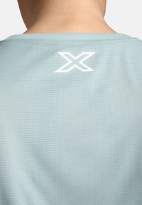 Světle modré sportovní tričko s malým bílým geometrickým logem na zadní straně, vyrobené z texturované látky s mřížkovým vzorem.