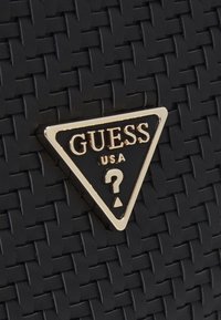 Czarna pleciona torba z wyraźnym trójkątnym złotym logo z napisem "GUESS USA" oraz wzorem znaku zapytania. Teksturowana powierzchnia, eleganckie akcenty.