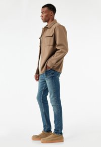 Chaqueta marrón de botones con dos bolsillos en el pecho, combinada con vaqueros ajustados azules y zapatos marrones claros. Textura suave, diseño casual.