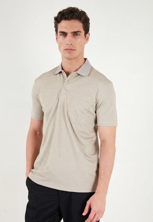 Uomo che indossa una polo beige a maniche corte con taschino sul petto e pantaloni neri, in piedi contro uno sfondo chiaro e semplice.