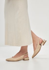 Beige slingback-kengät, joissa on pistemäinen kärki, nahkamateriaali, matala korko ja kultaiset yksityiskohdat korossa. Yhdistettynä vaalean väriseen hameeseen.