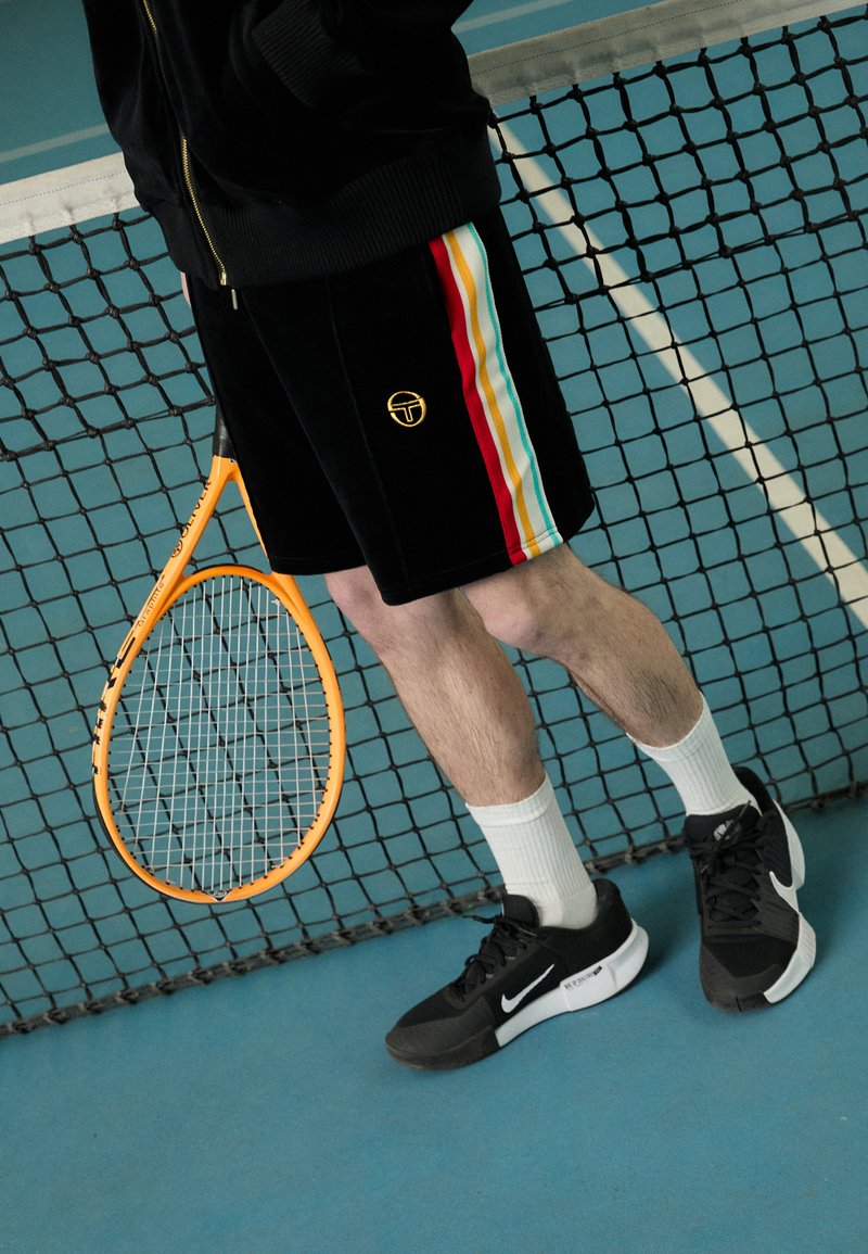 Mustad mustad lühikesed püksid, millel on mitmevärvilised külgrihmad, kuldne logo, kombineeritud mustade tenniste ja valgete sokkidega, hoides oranži tennisereketit.