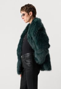 Teal fuskpälsjacka med en lurvig textur, bärs över en svart ribbad topp och svarta läderbyxor, med hög krage och öppen front.