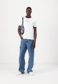 Camiseta blanca de manga corta con ribetes negros y el logo "GUESS JEANS", combinada con unos vaqueros holgados de color azul claro y zapatillas blancas. Diseño casual.