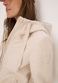 Veste en polaire beige avec capuche, dotée d'une fermeture éclair et d'un ruban portant l'inscription "FAIS TON PROPRE CHEMIN". Comprend un écusson logo sur la manche.
