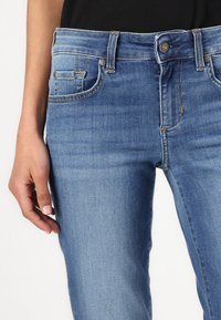 Modre jeans s srednjim pasom, zasnovo s petimi žepi in subtilnimi šivi na žepih. Tkanina se zdi mehka in teksturirana.
