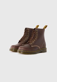 Botas de cuero marrón hasta el tobillo con costuras amarillas, punteras redondeadas, ojetes metálicos y tiradores. La suela es resistente y ligeramente elevada.