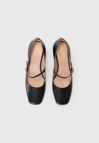 Black leather shoes with square toes, featuring crossed straps and a gold-tone buckle. The insole has a heart logo and light beige color.