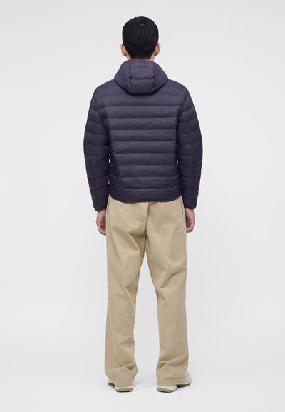 Homme portant une doudoune bleu marine foncé avec capuche et un pantalon beige ample, debout dos à l'objectif sur un fond blanc uni.