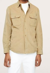 Mand iført en beigefarvet kordfløjl skjorte med knaplukning og to brystlommer, over en hvid t-shirt og hvide bukser.