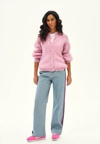 Žena s dlhými kučeravými vlasmi, oblečená v ružovom plyšovom kardigáne, modrých džínsoch s bočnými pruhmi a ružovými teniskami, stojaca pred jednoduchým pozadím.