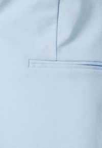 Tela azul claro con un bolsillo de ojal horizontal y detalle de costura vertical, mostrando una textura suave y costuras limpias.