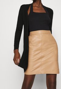 Cardigan nero sopra un top halter nero a coste, abbinato a una gonna in pelle beige a vita alta con dettagli nelle cuciture e una texture liscia.