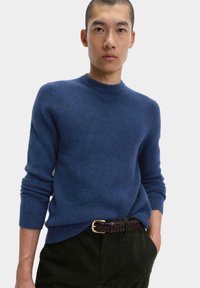 Modrý pletený svetr s kulatým výstřihem a žebrovanými manžetami, nošený přes tmavě zelené kordové kalhoty, vybavený pleteným hnědým opaskem.