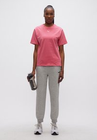 Γυναίκα όρθια που φοράει ροζ μπλουζάκι Puma, γκρι φόρμα Puma, λευκά αθλητικά παπούτσια, κρατώντας ένα μπουκάλι νερού Puma στο δεξί της χέρι.