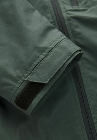 Manche de veste vert foncé avec sangle Velcro réglable et fermeture éclair noire visible sur fond de tissu.