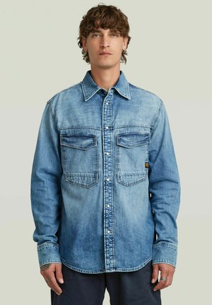 Hombre con cabello rizado que lleva una camisa de mezclilla azul con botones y dos bolsillos en el pecho, de pie frente a un fondo claro y liso.