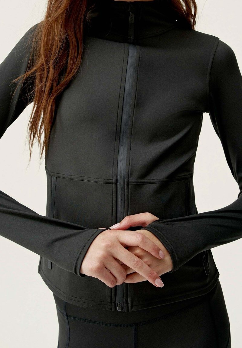 Svart träningsjacka med hög krage, framsida dragkedja, figursydda långa ärmar och slät textur. Har platta sömmar och en elegant design.