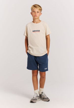 Beige katoenen t-shirt met blauwe en rode tekst, gecombineerd met marineblauwe shorts. Gestructureerde stof en eenvoudig ontwerp met zijzakken. Grijze sneakers.