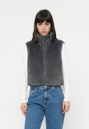 Grijze nepbont vest met hoge kraag, ritssluiting aan de voorkant en elastische onderrand, gedragen over een witte lange mouwen shirt, gecombineerd met blauwe jeans.