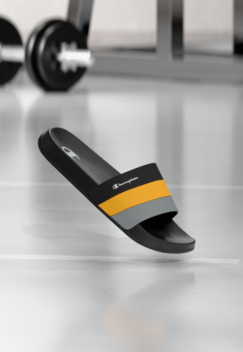 Sort, orange og grå Champion slide-sandal svæver over et fitnessgulv med slørede håndvægte i baggrunden.