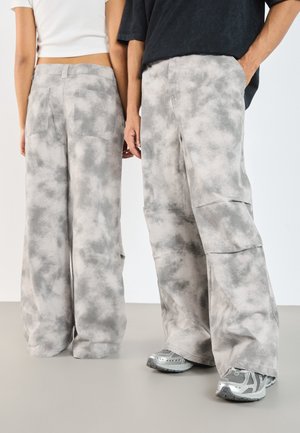 Dwie osoby noszące szerokie spodnie tie-dye w kolorach szarym i białym, jedna z czarną koszulką i sportowymi butami, druga z białym krótkim topem, stojące obok siebie.