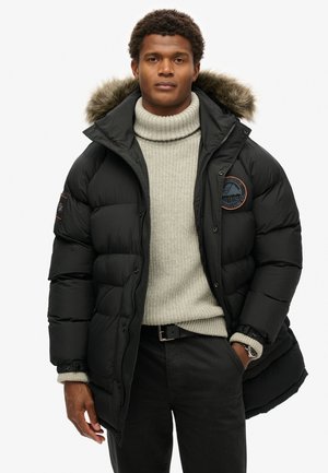 Chaqueta de plumas negra con capucha forrada de piel, cierre de cremallera y un parche redondo. Llevada sobre un suéter de cuello alto gris claro y pantalones negros.
