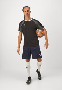Sort og grå sportstrøje med hvide accenter, blå shorts, hvide sokker og sorte fodboldstøvler. Personen holder en orange fodbold.