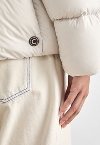 Puhasta kremna jakna z okroglo gumbasto oznako na rokavu, v kombinaciji s svetlo bež hlačami z modrim šivom na žepu.