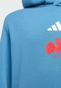 Blå hættebluse lavet af tekstureret stof med et rødt høne grafik og hvide stribede logo accenter på den øverste venstre side.