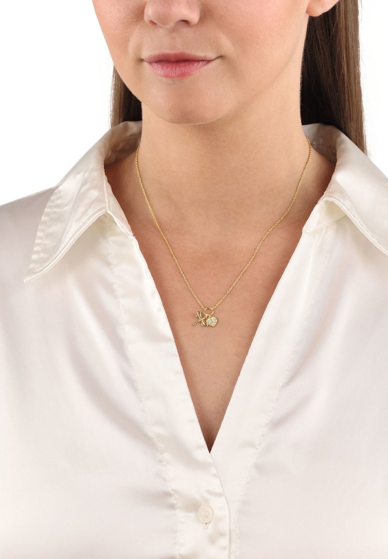 Kvinna som bär ett guldhalsband med libellor och hjärtformade kristallberlocker på en kedja, tillsammans med en vit satinblus.