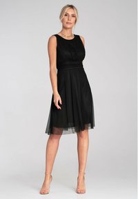 Schwarzes ärmelloses Kleid mit einem figurbetonten Oberteil und einem fließenden Tüllrock. Hat einen runden Ausschnitt und eine Taillenbetonung.