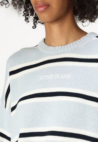 Hellblauer gestreifter Strickpullover mit breiten navyblauen und weißen Streifen, ausgestattet mit einem gerippten Ausschnitt und gesticktem "GUESS JEANS"-Logo auf der Vorderseite.