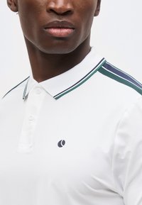 Κοντινό ενός άνδρα που φοράει λευκό πόλο μπλουζάκι με γιακά και λεπτομέρειες ώμων σε πράσινη και ναυτικό ρίγα, καθώς και μικρό μαύρο λογότυπο στο στήθος.