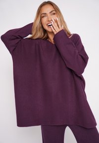 Übergroßer lila Strickpullover mit langen Ärmeln und weitem Ausschnitt, der eine glatte Textur aufweist. Kombiniert mit passenden lila Hosen.