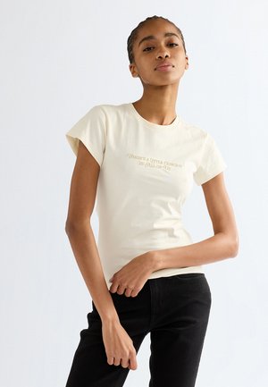 Jeune femme portant un t-shirt crème ajusté avec le texte "There's a little cowboy in all of us" et un pantalon noir, posant devant un fond uni.