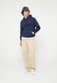 Ναυτικό μπλε φούτερ με κουκούλα και μπροστινή τσέπη, με ανάγλυφο λογότυπο, σε συνδυασμό με μπεζ παντελόνι και λευκά αθλητικά παπούτσια. Μοντέλο που φοράει σκούρο καπέλο.