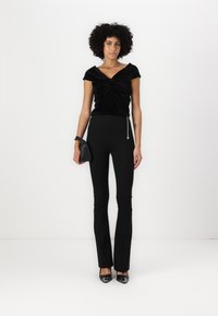 Top de terciopelo negro con un escote torcido, combinado con pantalones negros ajustados que tienen una cremallera lateral. Los accesorios incluyen un pequeño bolso negro.