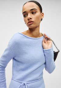 Pull bleu clair en matière douce, avec un décolleté asymétrique, des manches longues et un détail de taille à cordon.