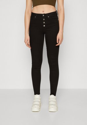 Kvinde iført højtaljede sorte skinny jeans med fire synlige knapper og hvide sneakers, stående mod en ensartet lys baggrund.