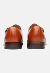 Scarpe in pelle marrone con finitura liscia, punta rotonda, doppia fibbia e tacco in legno sovrapposto. Il design presenta linee classiche e pulite.
