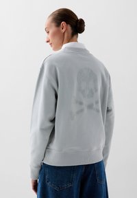 Grijze sweatshirt met een gestructureerd skull-and-crossbones ontwerp op de achterkant, voorzien van een relaxte pasvorm en geribbelde manchetten en zoom.