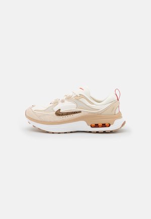 Beżowo-biały sneaker Nike Air Max z brązowym logo, pomarańczową poduszką powietrzną i czerwoną pętelką na białym tle.