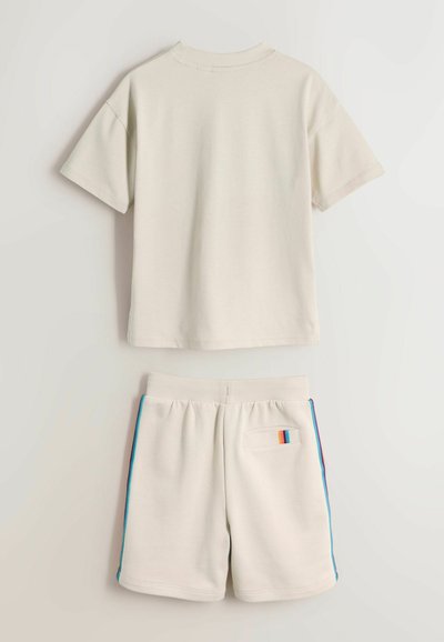 Beige kortærmet skjorte og matchende shorts med elastisk talje, blå og orange sidestriber samt en lille baglomme med regnbuedetalje.