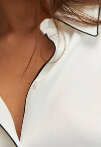 Hvid skjorte med en klassisk krave med sort piping, knaplukning ved halsen og en glat, blød tekstur.