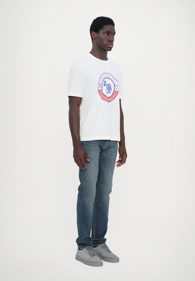 Tricou alb din bumbac cu un grafic circular care prezintă o zebră, asociat cu blugi denim albaștri și adidași gri.
