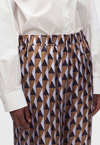 Personne portant une jupe à motifs géométriques marron, noire et blanche avec une taille élastique et une chemise blanche à manches longues boutonnée.