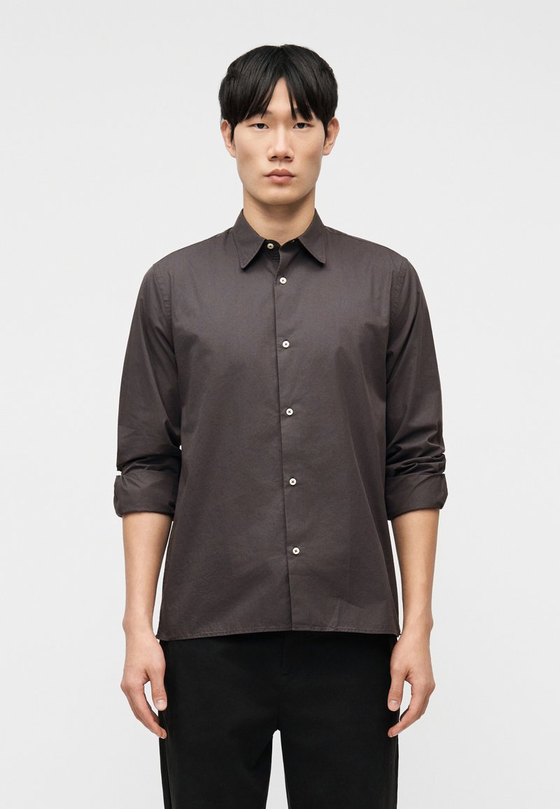 Νεαρός άνδρας όρθιος, φορούσε σκούρο γκρι πουκάμισο με κουμπιά και μανίκια γυρισμένα, καθώς και μαύρο παντελόνι, κοιτώντας ευθεία μπροστά σε λευκό φόντο.