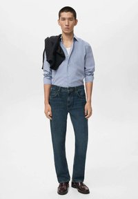 Gestreepte blauwe button-up shirt, donkere denim jeans, zwarte trui over de schouder, donkerbruine schoenen, staand op een effen achtergrond.