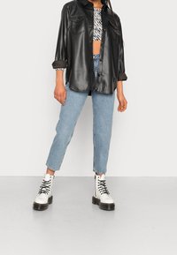 Zwarte faux leren overhemdjack met twee borstzakken over een gestreept crop top, gecombineerd met lichtblauwe straight-leg jeans en witte veterschoenen.